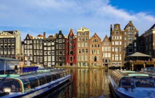 Colorful historic buildings along Amsterdam canals with boats in the foreground, reflecting in calm water.