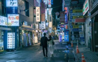 An Asian couple walks through the vibrant, neon-lit streets of Seoul at night, surrounded by shops and signs.