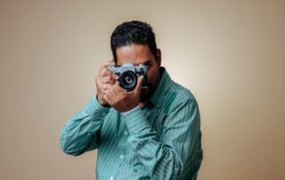 Man posing with a vintage camera in a studio setting wearing a striped green shirt.
