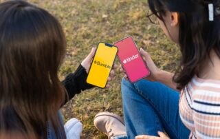 Two women using Bumble and Tinder apps on smartphones in a park setting.