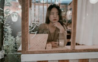 A young woman sitting thoughtfully at a cafe, gazing through the window with plants around her.