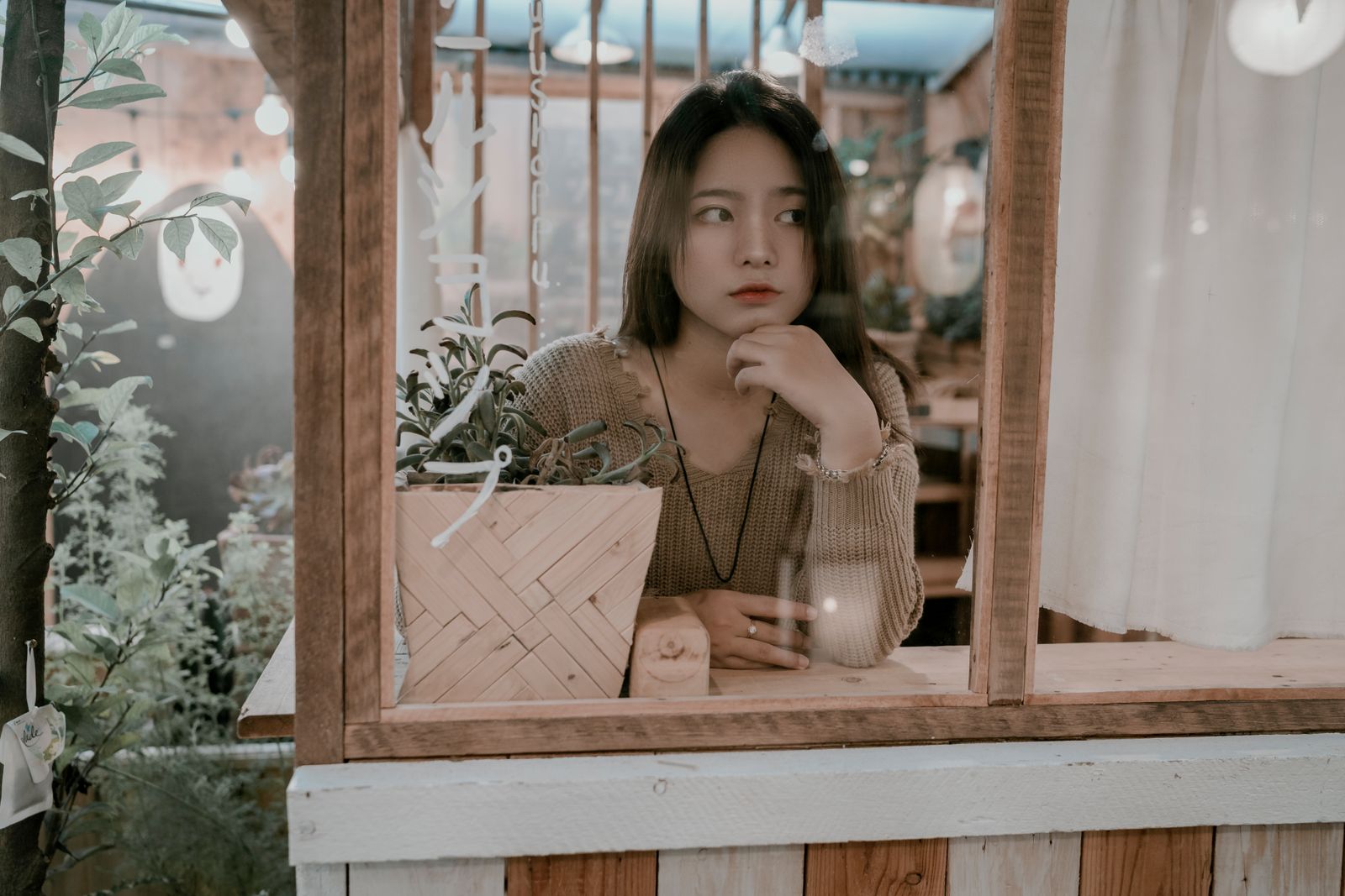 A young woman sitting thoughtfully at a cafe, gazing through the window with plants around her.