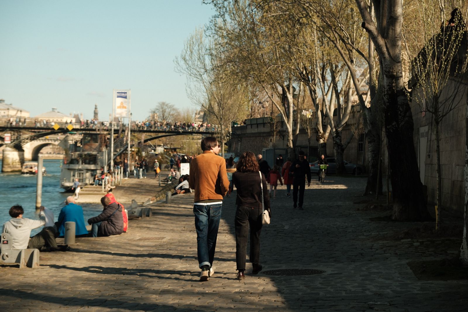 A couple walks hand in hand by the Seine in Paris, surrounded by spring ambiance and city life.