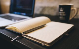 Open notebook with a pen on a wooden desk beside a laptop, ideal for blogging and work-related themes.