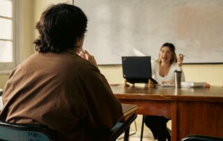 Student and teacher engage in discussion in a classroom setting with a laptop and whiteboard visible.