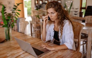 A woman in a bright room using a laptop and phone, blending work with home comfort.