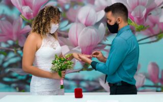 A couple exchange rings wearing masks during an intimate wedding with floral backdrop.