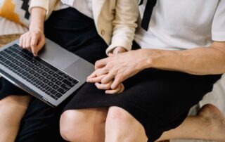 A close-up of a couple sitting together, using a laptop while holding hands.