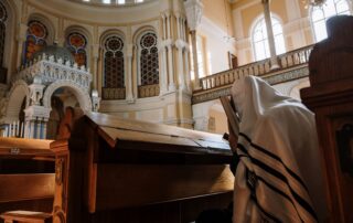 A person wearing a tallit engaged in prayer inside a beautifully designed synagogue with stained glass windows.