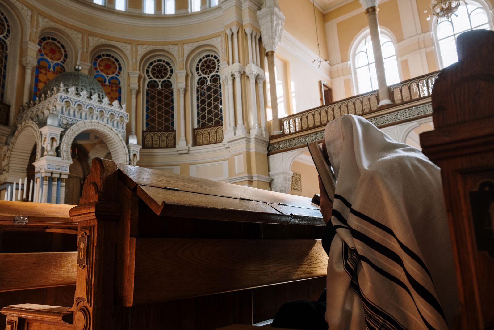 A person wearing a tallit engaged in prayer inside a beautifully designed synagogue with stained glass windows.