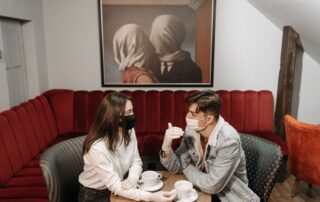 A couple enjoys coffee while wearing masks in a cozy café, highlighting the pandemic's impact.