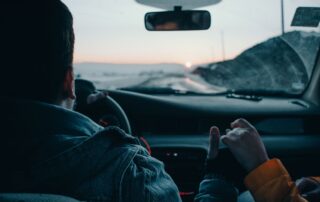 Back view of crop anonymous couple of travelers driving modern automobile along asphalt road at sunset