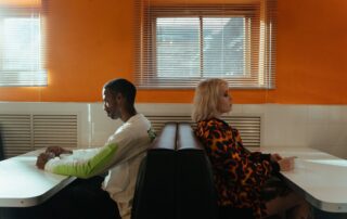 A couple sits back-to-back in a diner, reflecting a tense moment or disagreement.