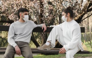 Young couple wearing masks sitting on a park bench, engaged in conversation, showcasing new normal.