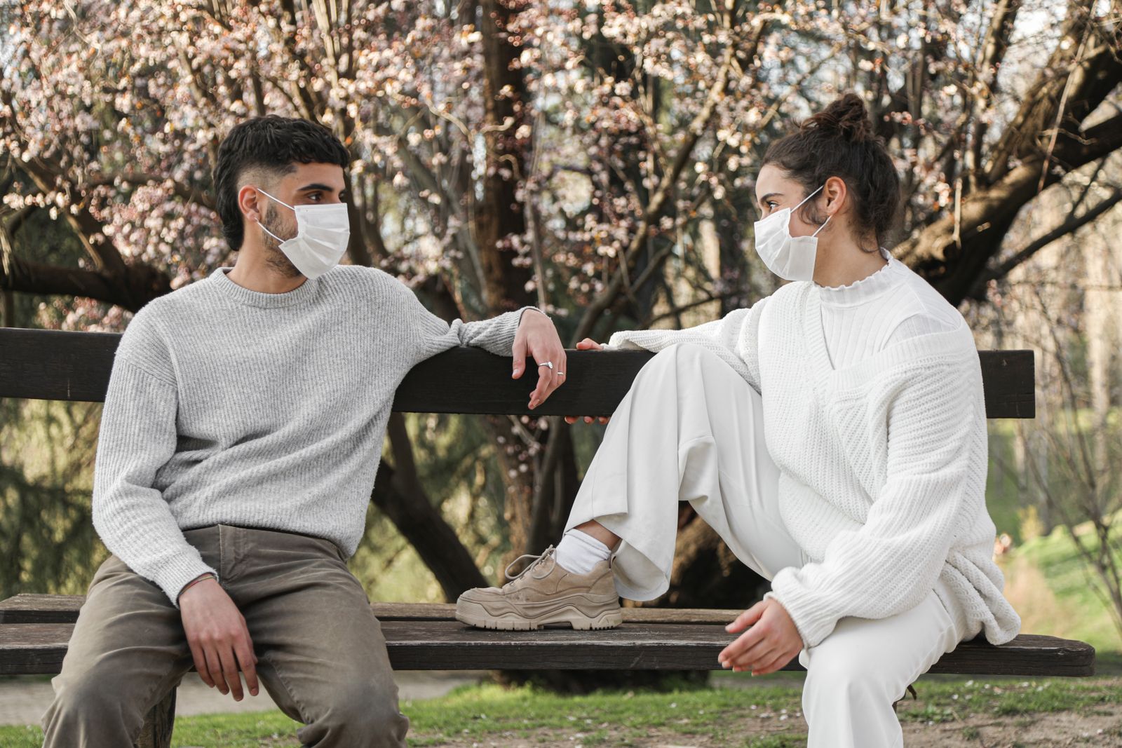 Young couple wearing masks sitting on a park bench, engaged in conversation, showcasing new normal.