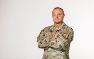 Military veteran standing with pride in camouflage uniform against a white background.