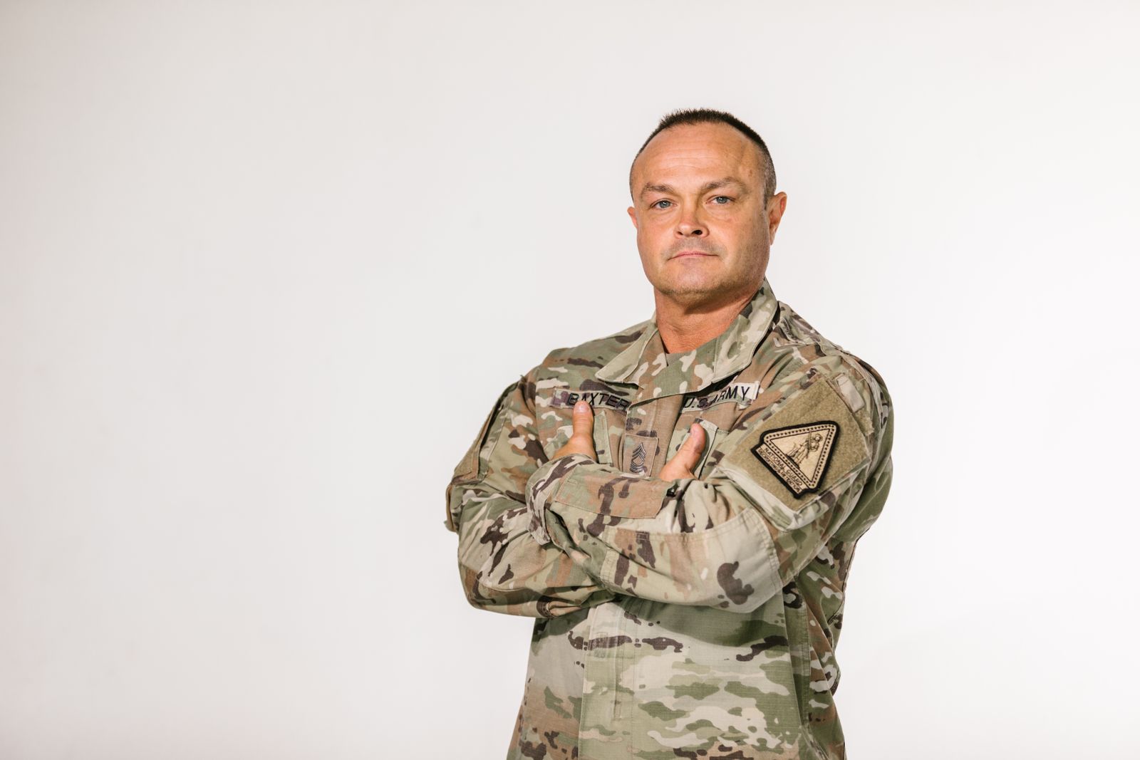 Military veteran standing with pride in camouflage uniform against a white background.