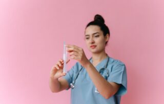 Female medical practitioner holding a syringe, standing in front of a pink background.