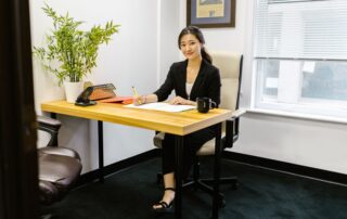 An Asian woman in business attire writing at a desk in a modern office setting.