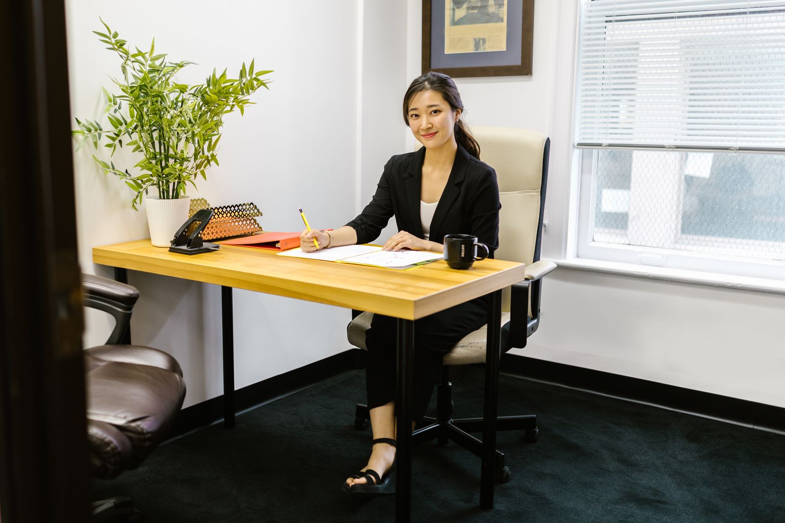 An Asian woman in business attire writing at a desk in a modern office setting.