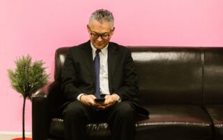 Asian man in a suit using smartphone, sitting on a leather sofa against a pink background.