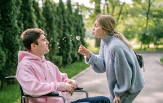 A woman blows a dandelion while a man in a wheelchair smiles, enjoying a sunny day outdoors.