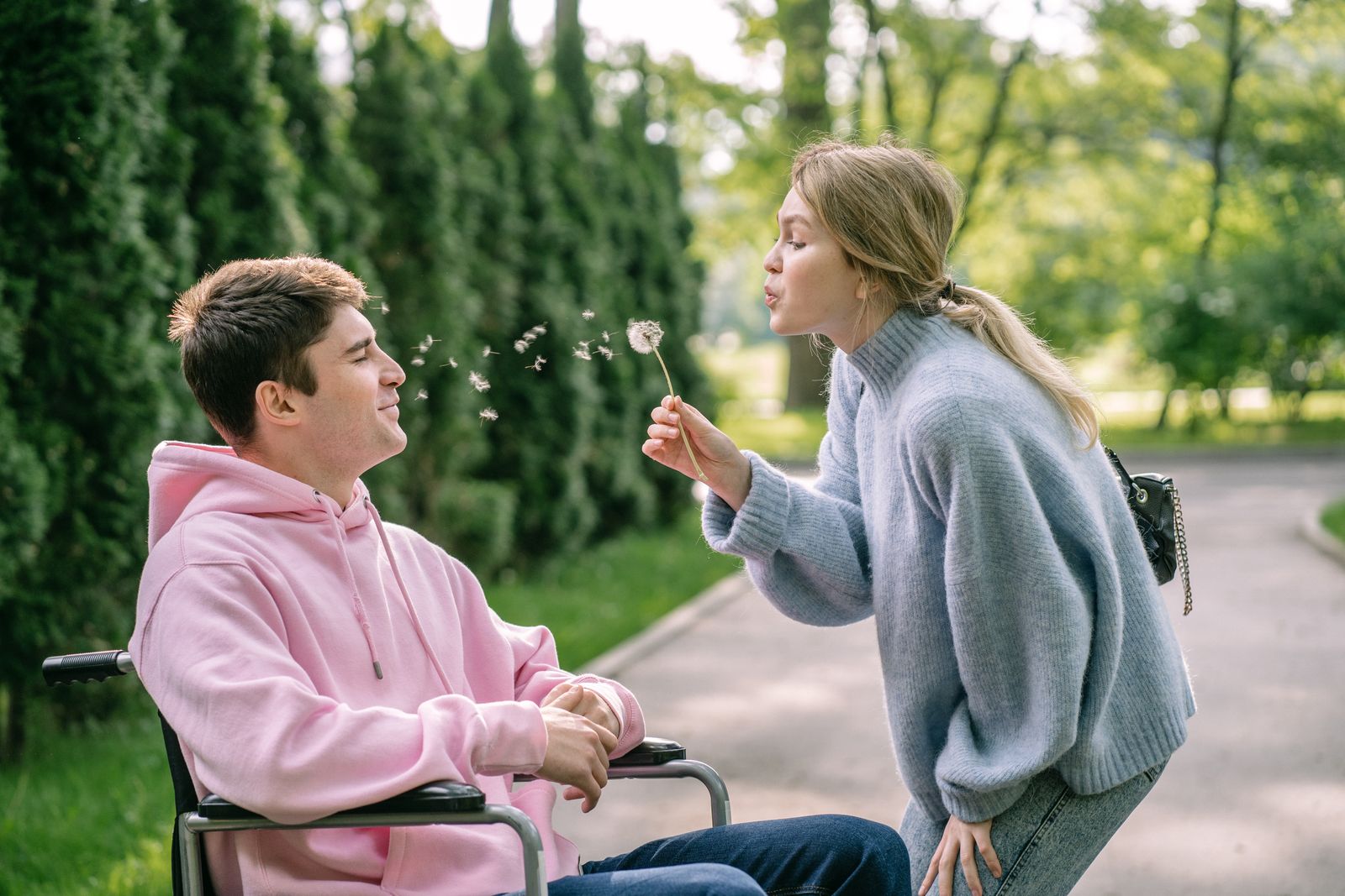 A woman blows a dandelion while a man in a wheelchair smiles, enjoying a sunny day outdoors.