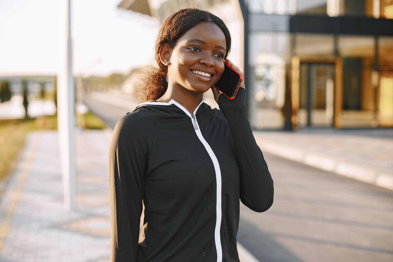 Young woman in sportswear smiling while talking on her smartphone outdoors.