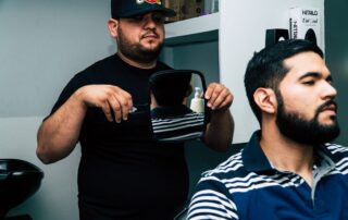 A barber in a cap shows a mirror to a client with a styled beard in a modern barbershop.